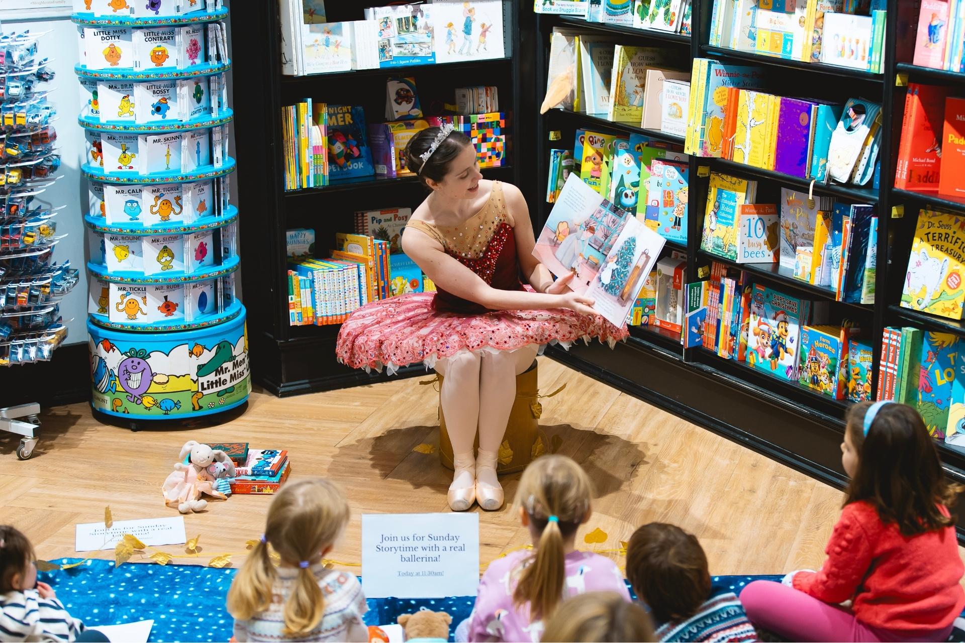 In a bookstore, a ballerina dressed in a pink tutu, pointe shoes and a tiara sits in front of a group of children reading a picture book of The Nutcracker.