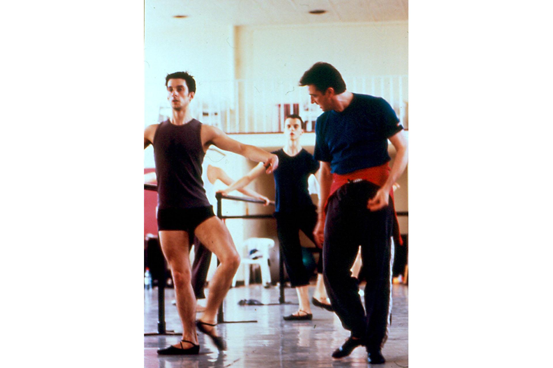 A dancer in black attire and black ballet shoes stands by the barre. The instructor stand by his side, looking at his body placement.