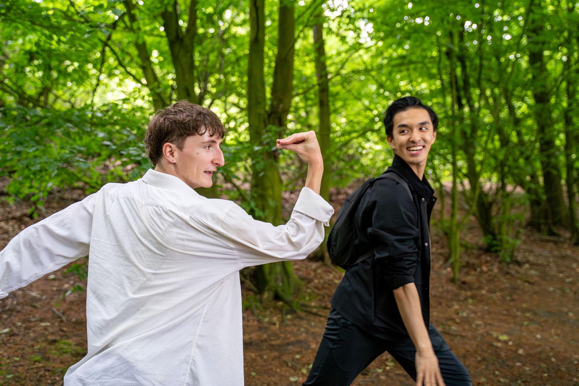 Two dancers stood in a woodland forest, one in a white shirt in the forefron with arm bent and palm closed to their face like a bird. The other dancer in black looks on smiling.