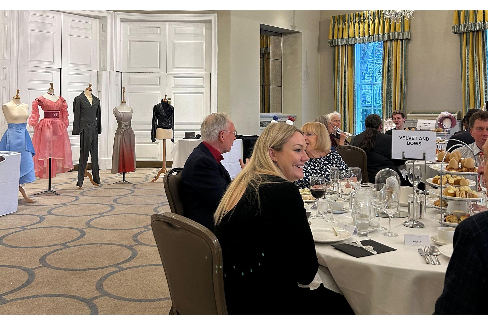 People sat at round tables with cacke stads filled with afternoon tea treats. In the back ground manaquins of costumes from Gentleman Jack.