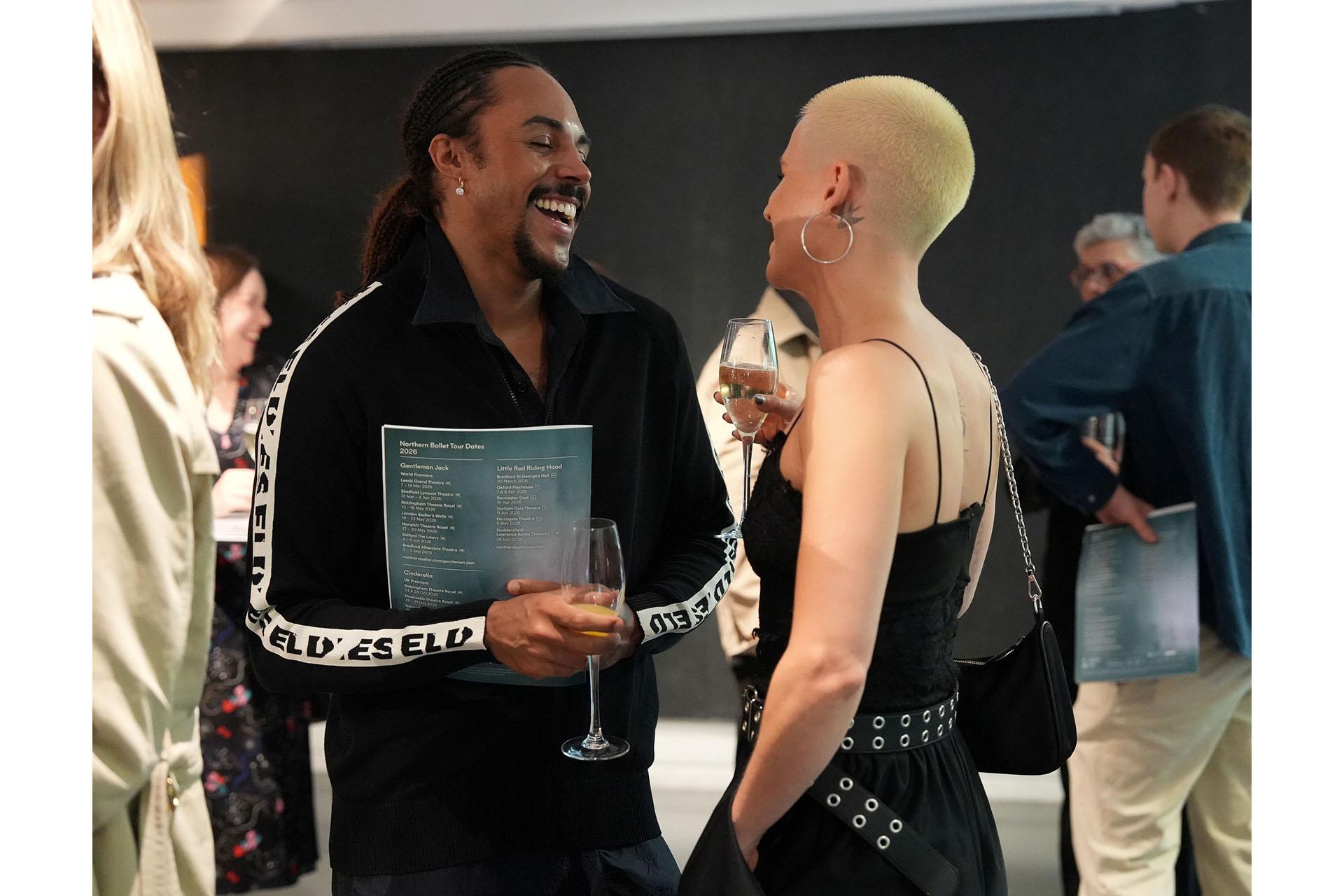 Two people talking, each holding a wine glass; the man smiles with an open mouth.