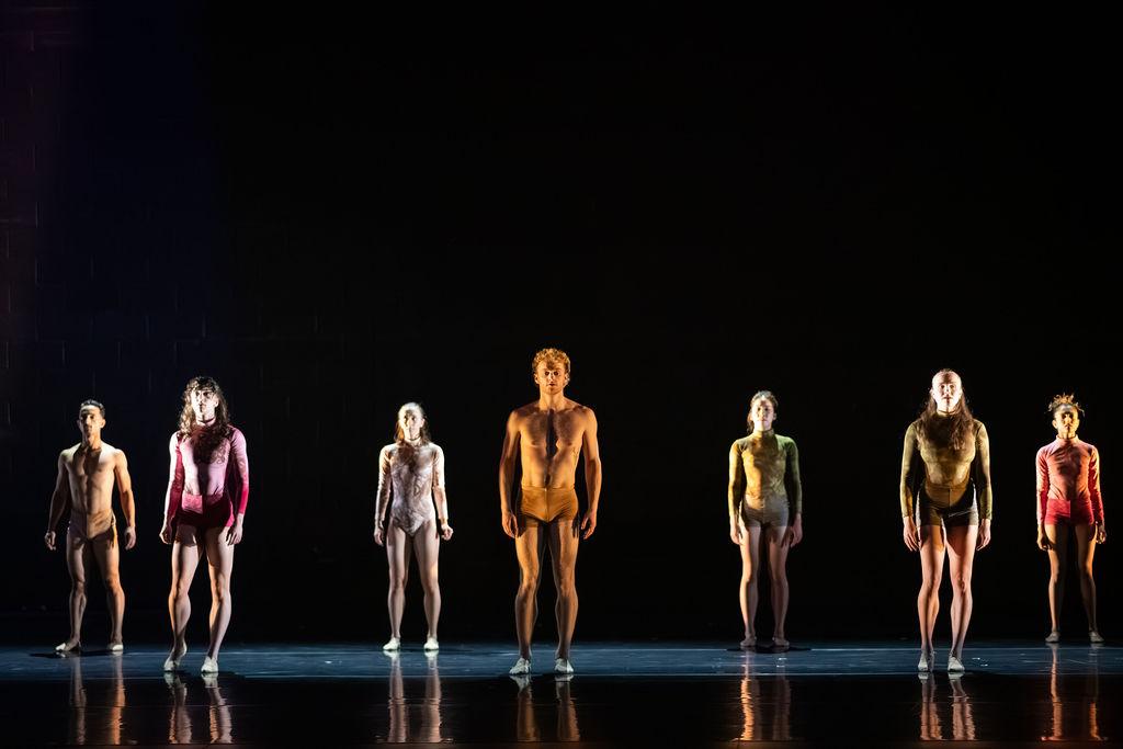Seven dancers stand on a black stage. They are wearing little clothing - the men are shirtless with small shorts on and the woman wear tight leotards. They are stood in a staggered line with their feet together, arms down at their sides and they are looking out, straight forward.
