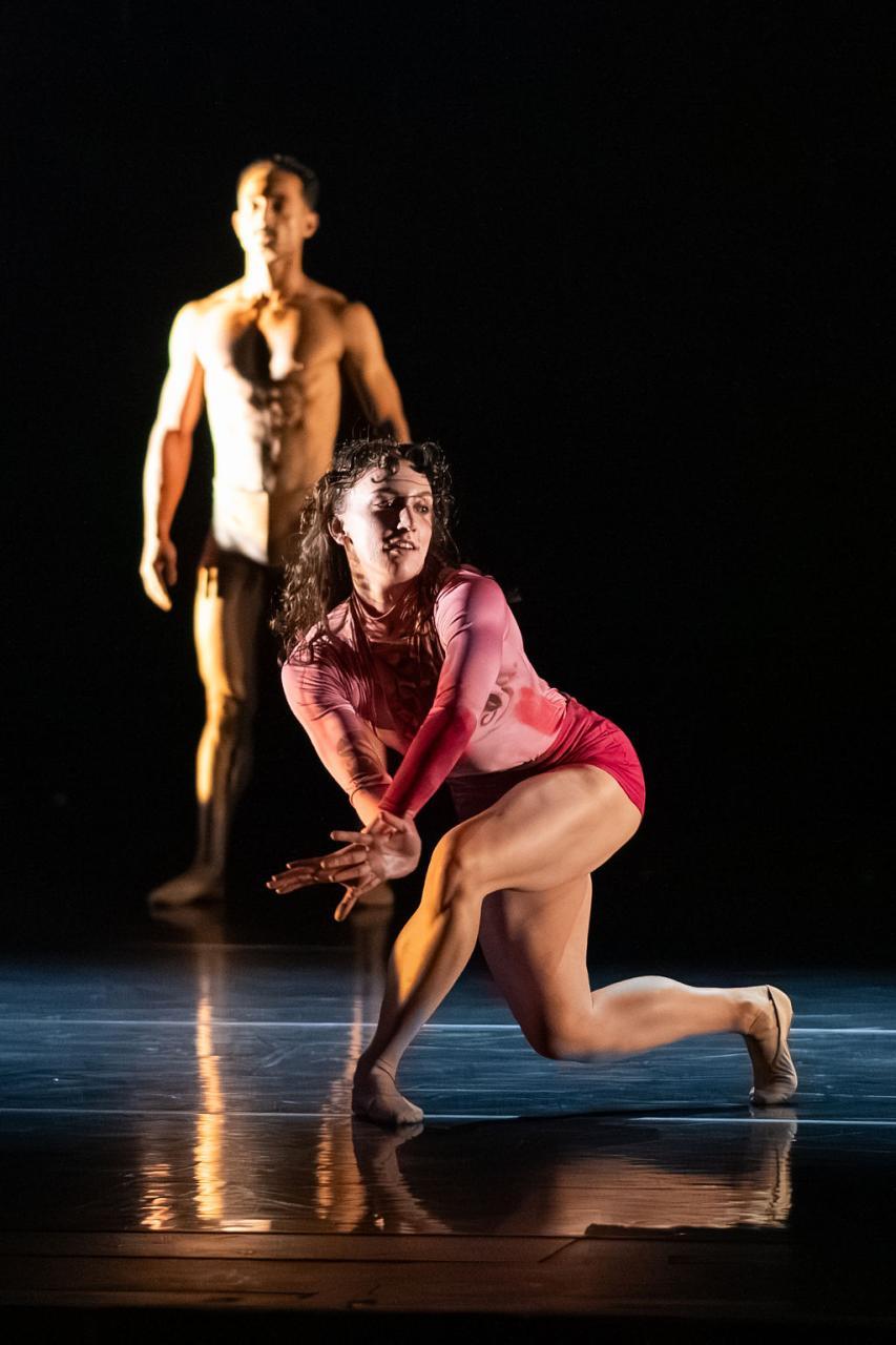 A female dancers in a pink long-sleeved leotard is pictured. She is in a lunge position with her left leg back. Her arms are out in front of her, crossed at the wrists. She looks to her left. There is also a male dancer in the background, stood still.