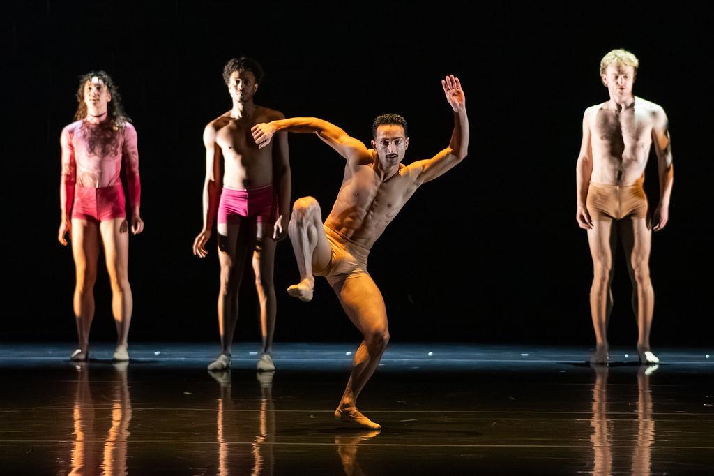 A male dancer is shirtless and in skin-coloured shorts. He balances on his left leg with his right up to his side, bent at the knee. His arms are up in the air, creating a curved silhouette. There are also three other dancers, one female and two male, pictured in the background and they are stood still, looking out.