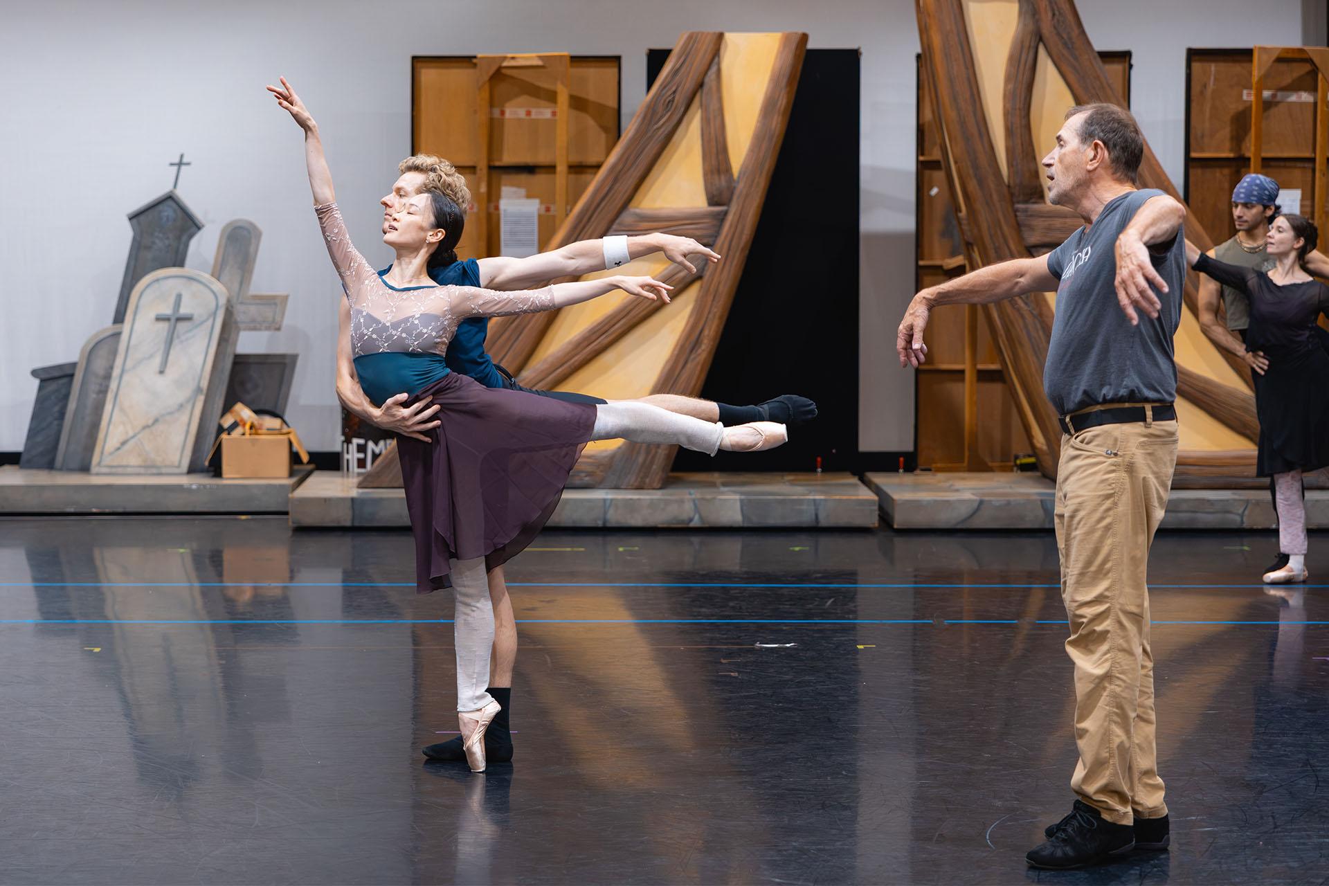 In the rehearsal room the choreographer watches closely as two dancers stand arabesque, in front she is en point and one arm lifted above her head, the other dancer imnmediately next to her performing the same move only with an arm around her for mutual support