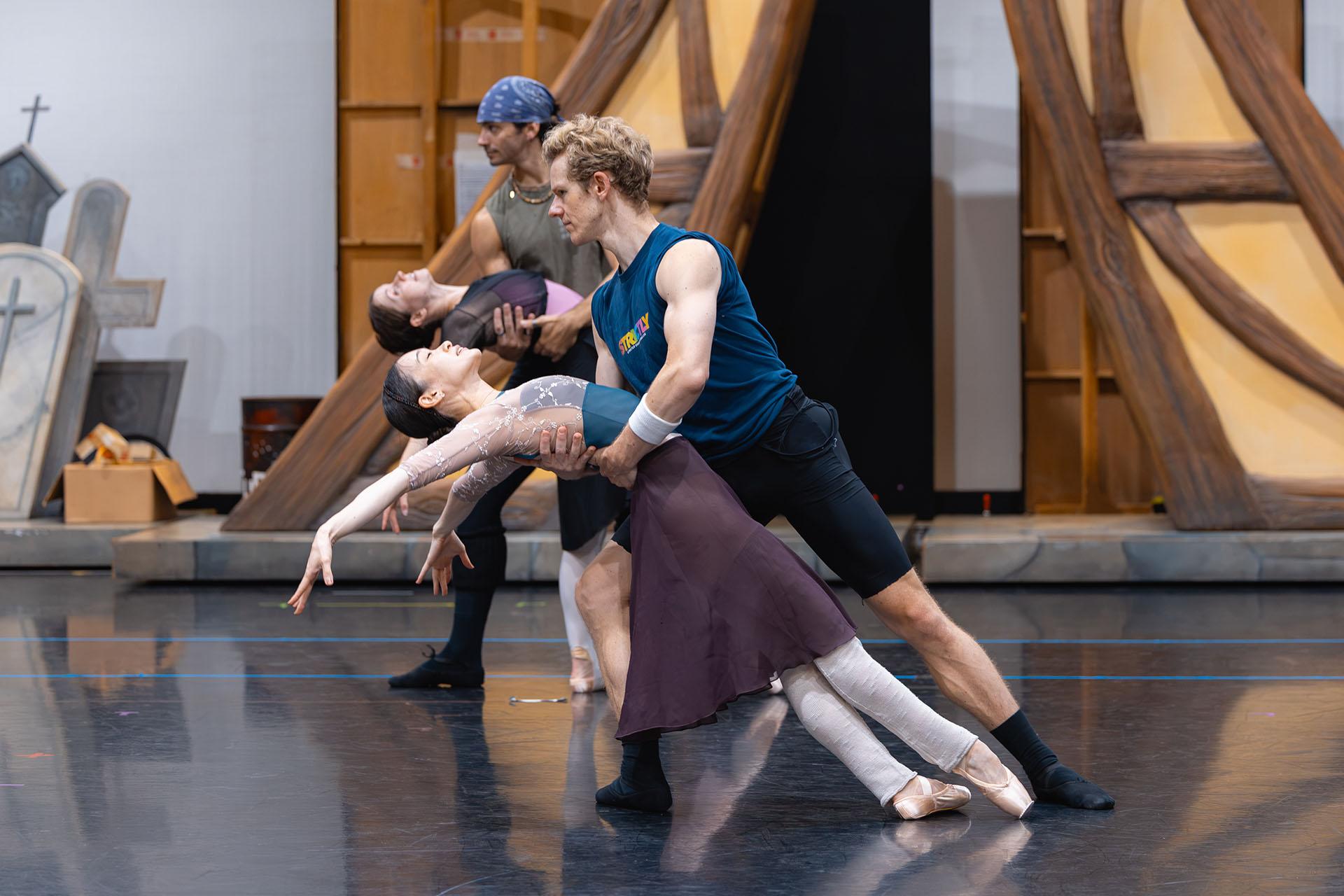 In rehearsal a woman dancer leans back, her legs forward, her only support another dancer holding her as she arches her back