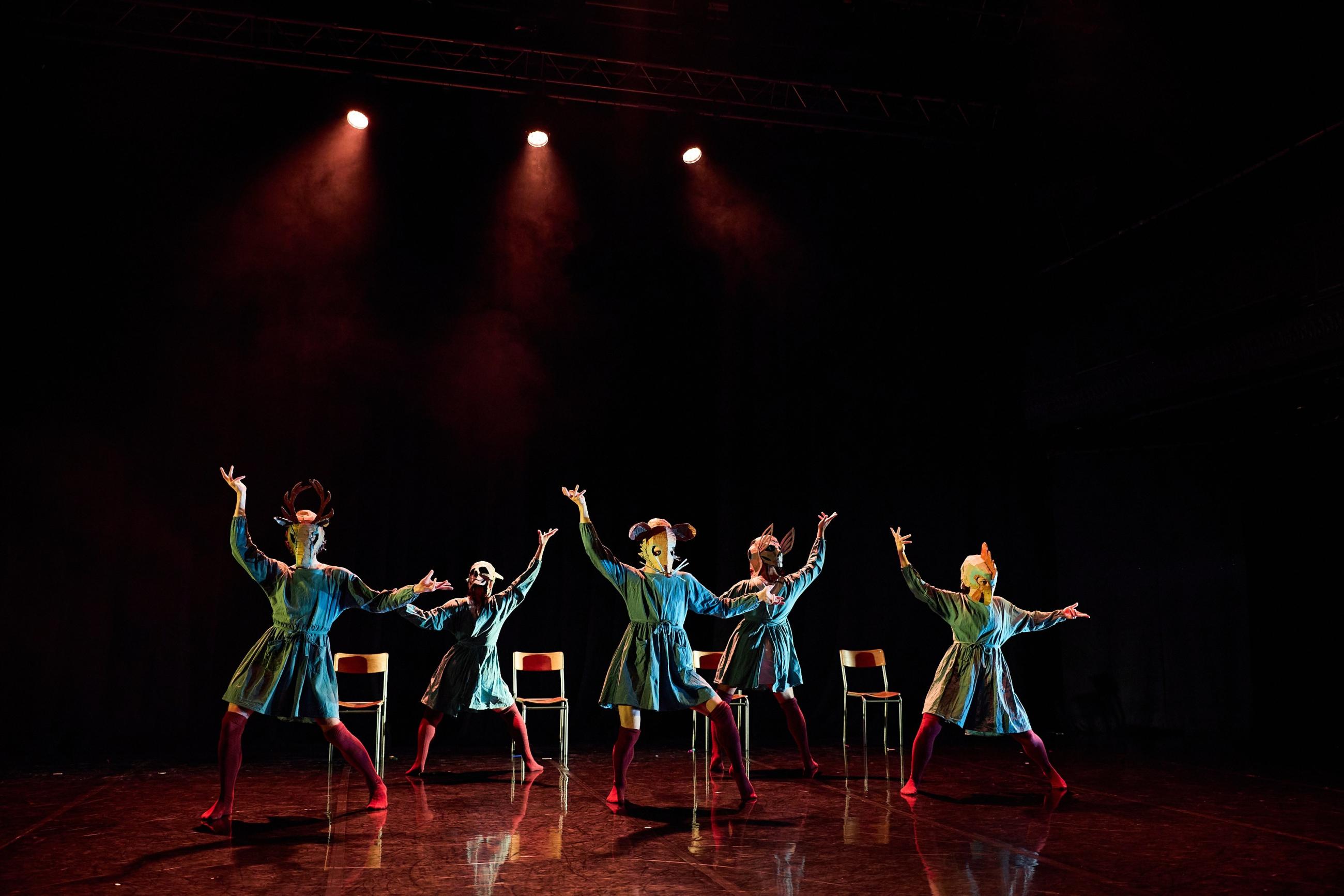 A group of 5 dancers with animal masks on (mouse, chicken, moose etc) stand in formation in front of a row of chairs. They are in various poses with their arms up around their heads.
