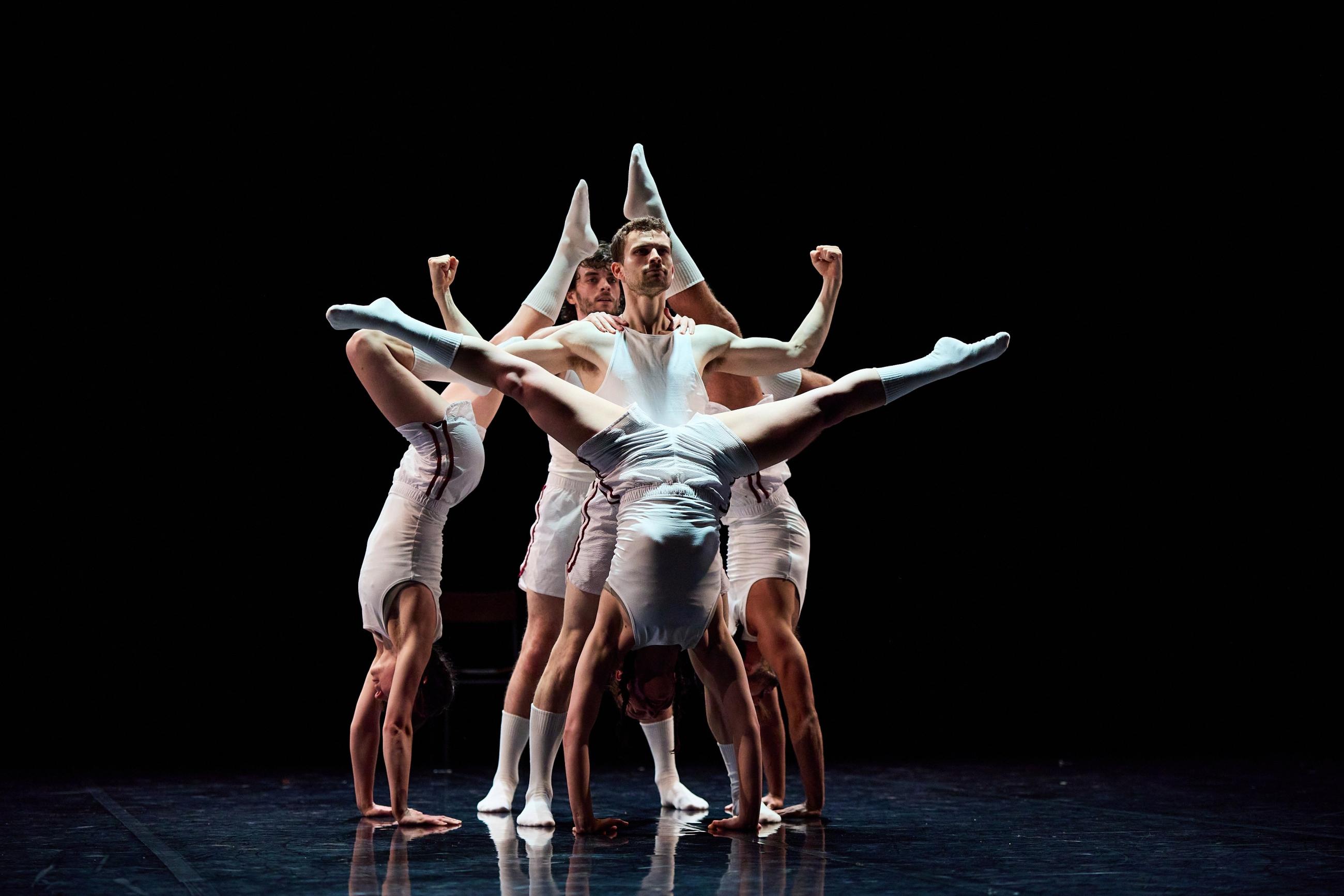 A group of 5 dancers all dressed in white are in formation. One man stands in the centre looking out, whilst two dancers are either side of him, doing handstands with their feet pointed. One dancer does a handstand in front of him, with their legs apart in a Y shape. One man stands behind to support the other dancers.