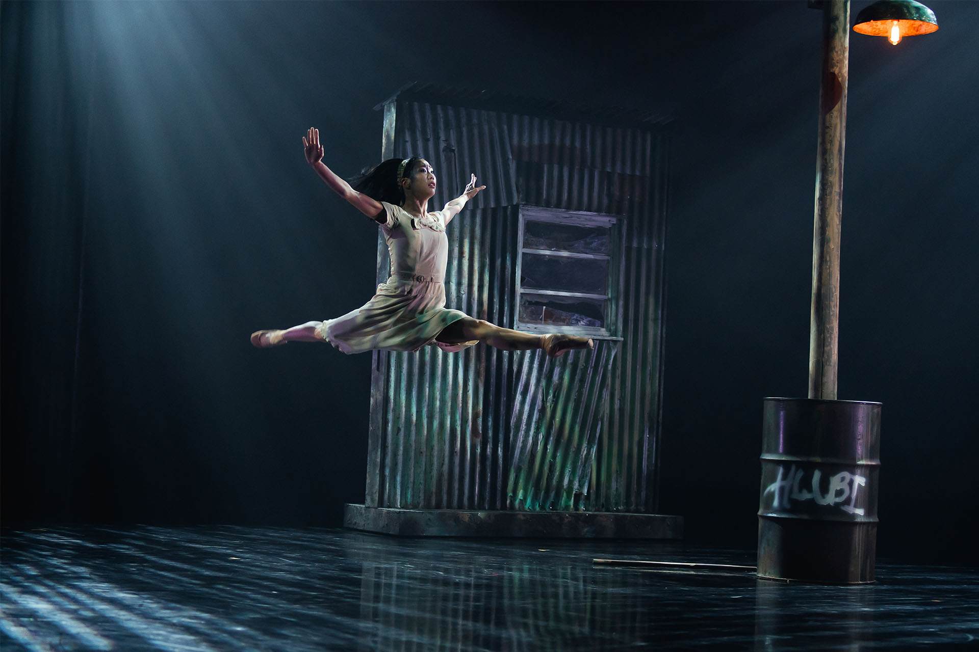 Leaping across the stage in atmospheric lighting, a shack of corrugated metal behind her, the dancer performs a graceful grand jete