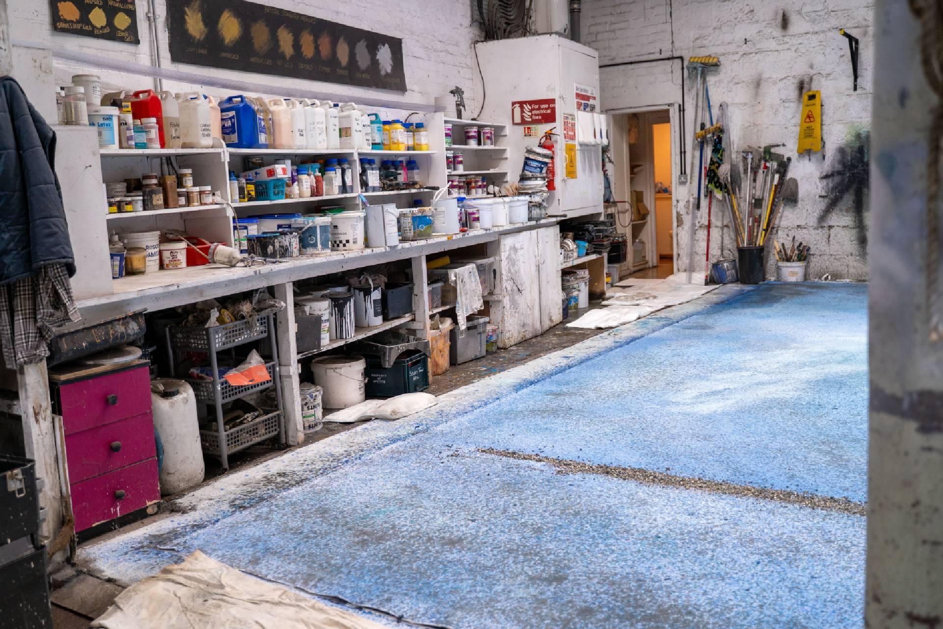 In a workshop, a large canvas lies on the floor with different shades of blue paint. Various bottles and tools are sitting on shelves on the wall.