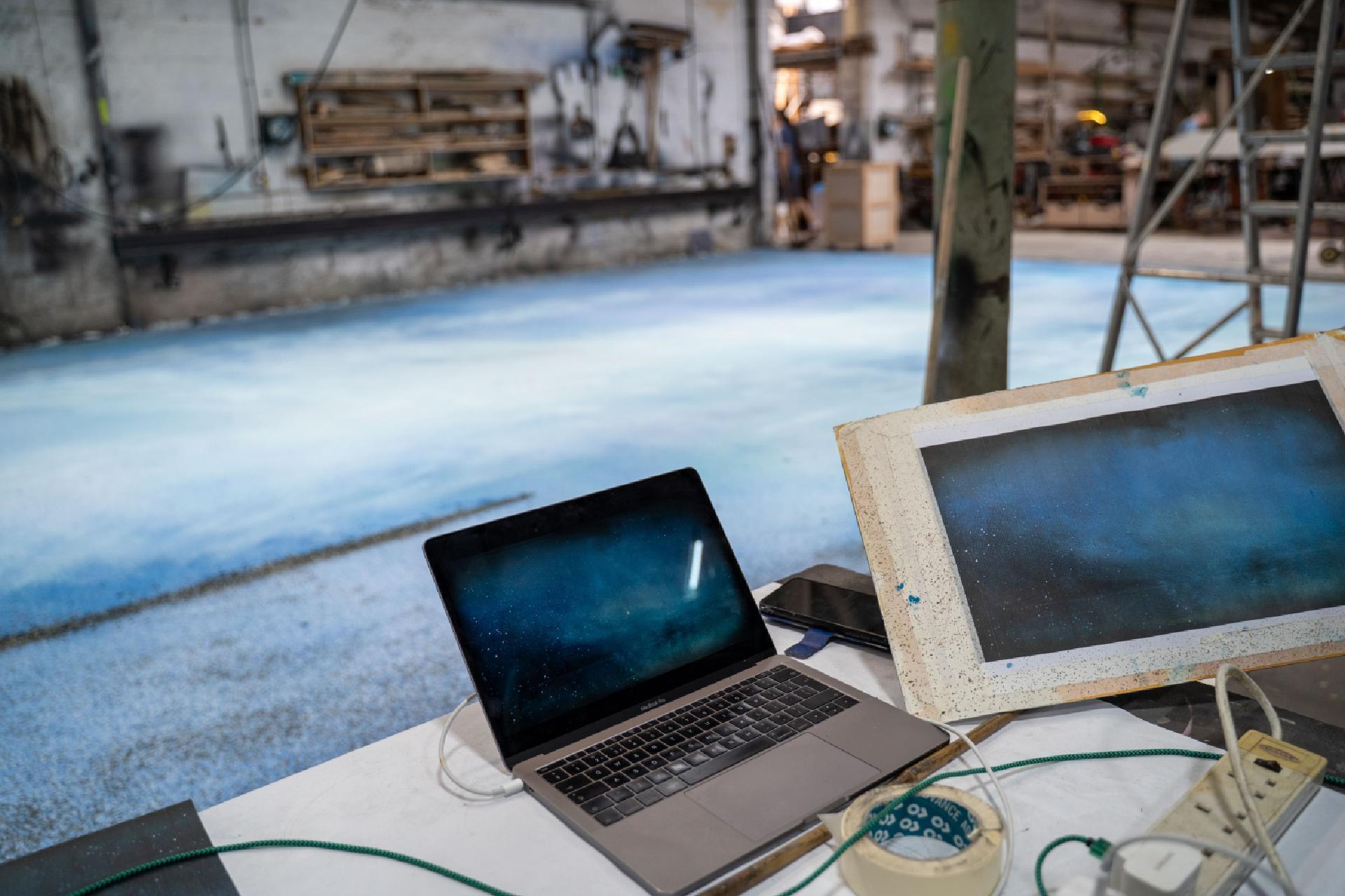 On a table, a laptop and a piece of paper show a print of a starry sky. In the background, a large canvas is on the floor, resembling the design of the print.