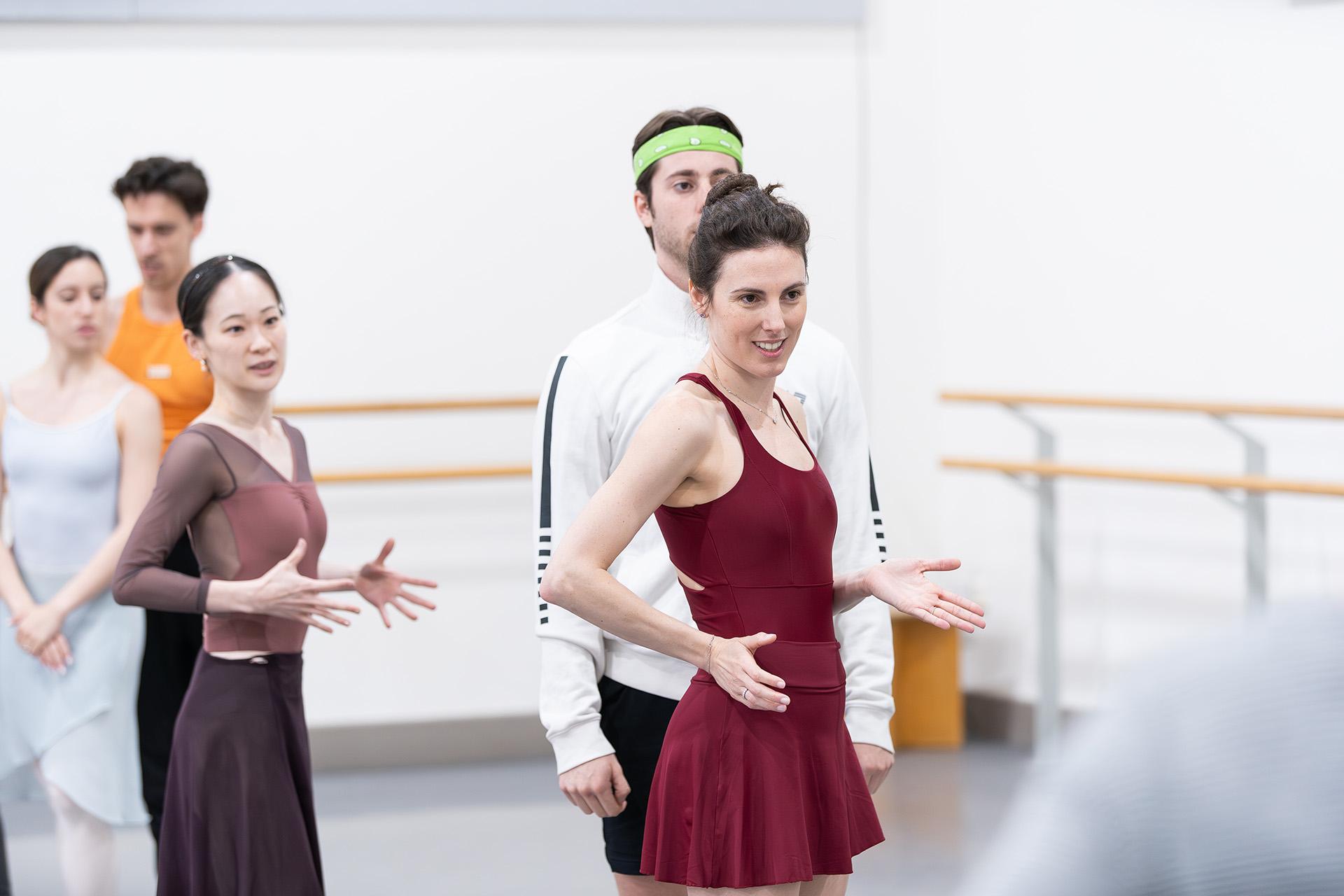 Choreographer in a red top and skirt demonstrates movement to a watching dancer who copies her hand positions