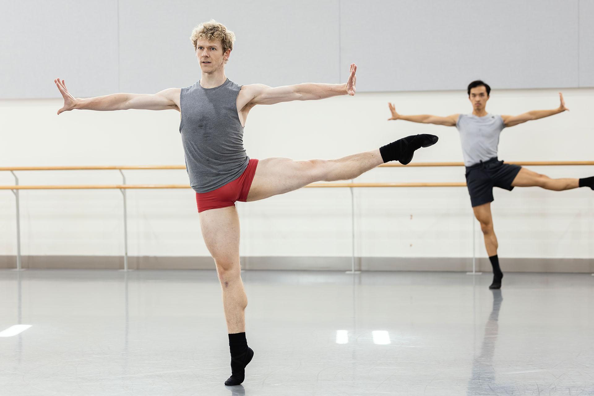 Male dancers performing the same move in rehearsals, holding their arms out, pals and fingers vertical, one leg on the floor, on their toes, the other sideways from their body, toes pointed