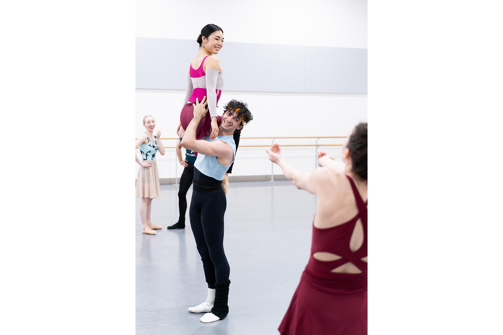 The choreographer looks on and advises the dancers, one hold the other on his shoulder, both smiling at the choreographer