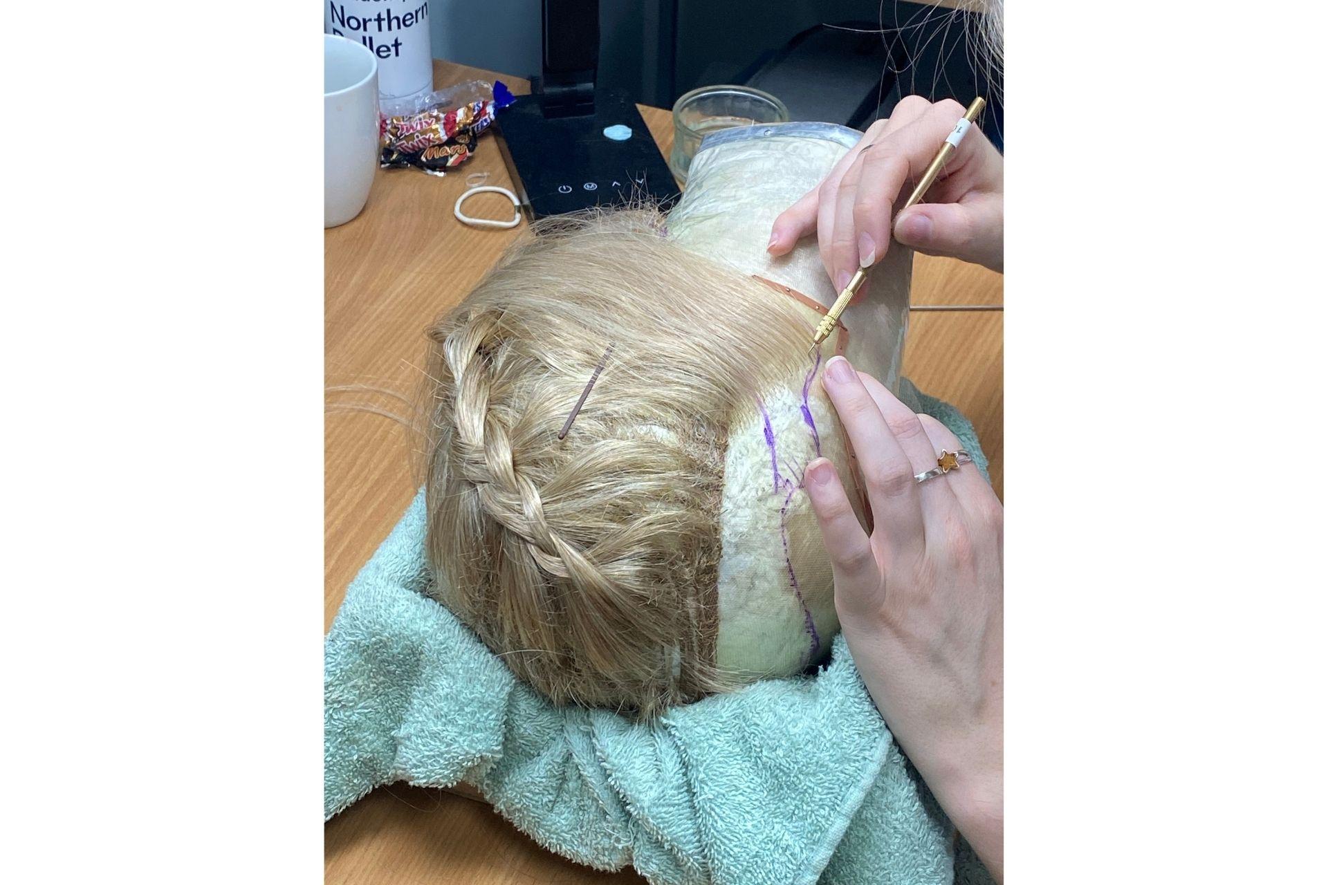 A mannequin head with a blonde wig is laid atop a table while a pair of hands works on forming the hairline