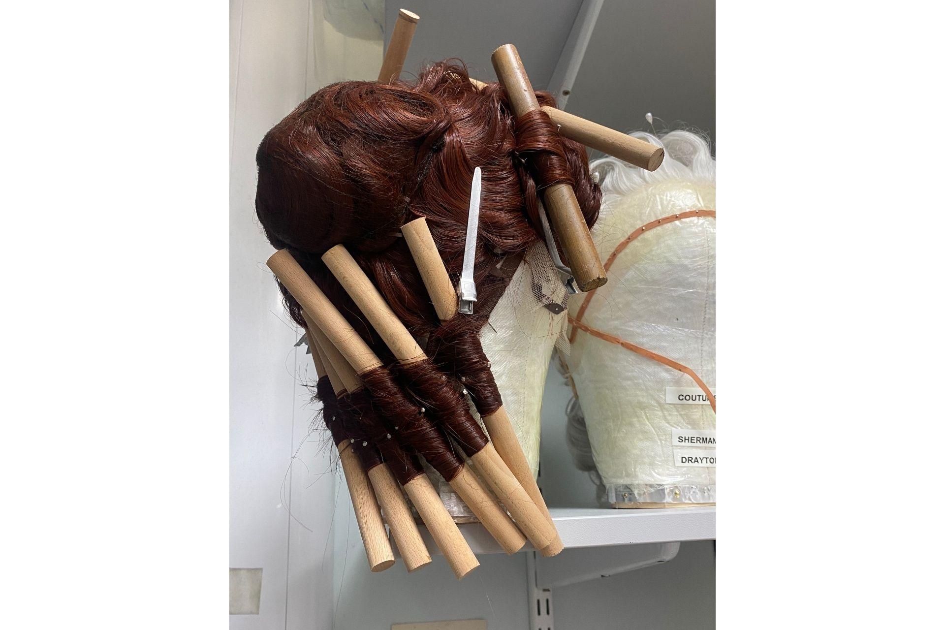 An intricately styled brown wig sits on a mannequin head with brown rolls of paper being used as curlers