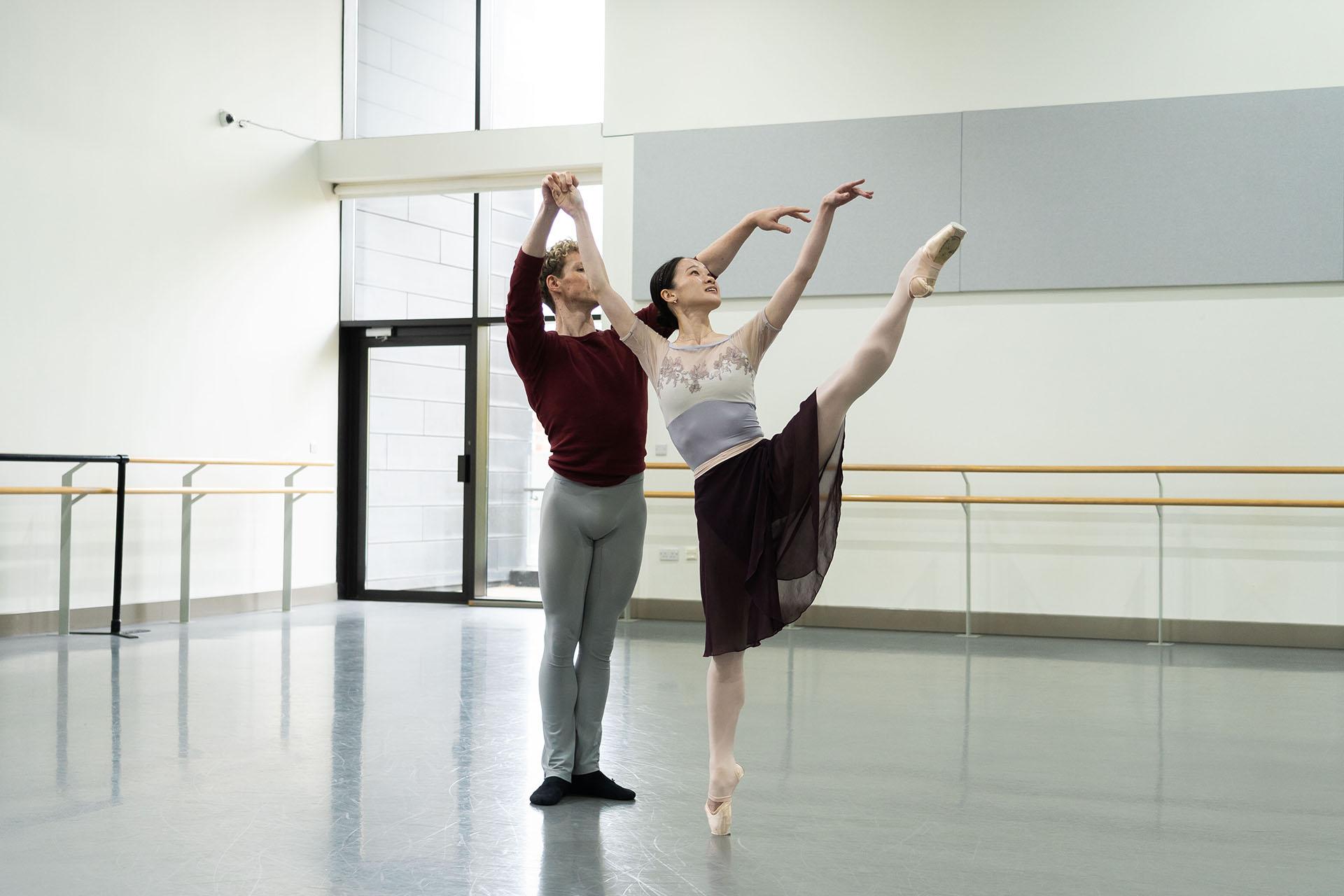 a dancer en pointe on one foot, the other leg raised in the air, her arms above her and one hand held for support by another dancer just behind her