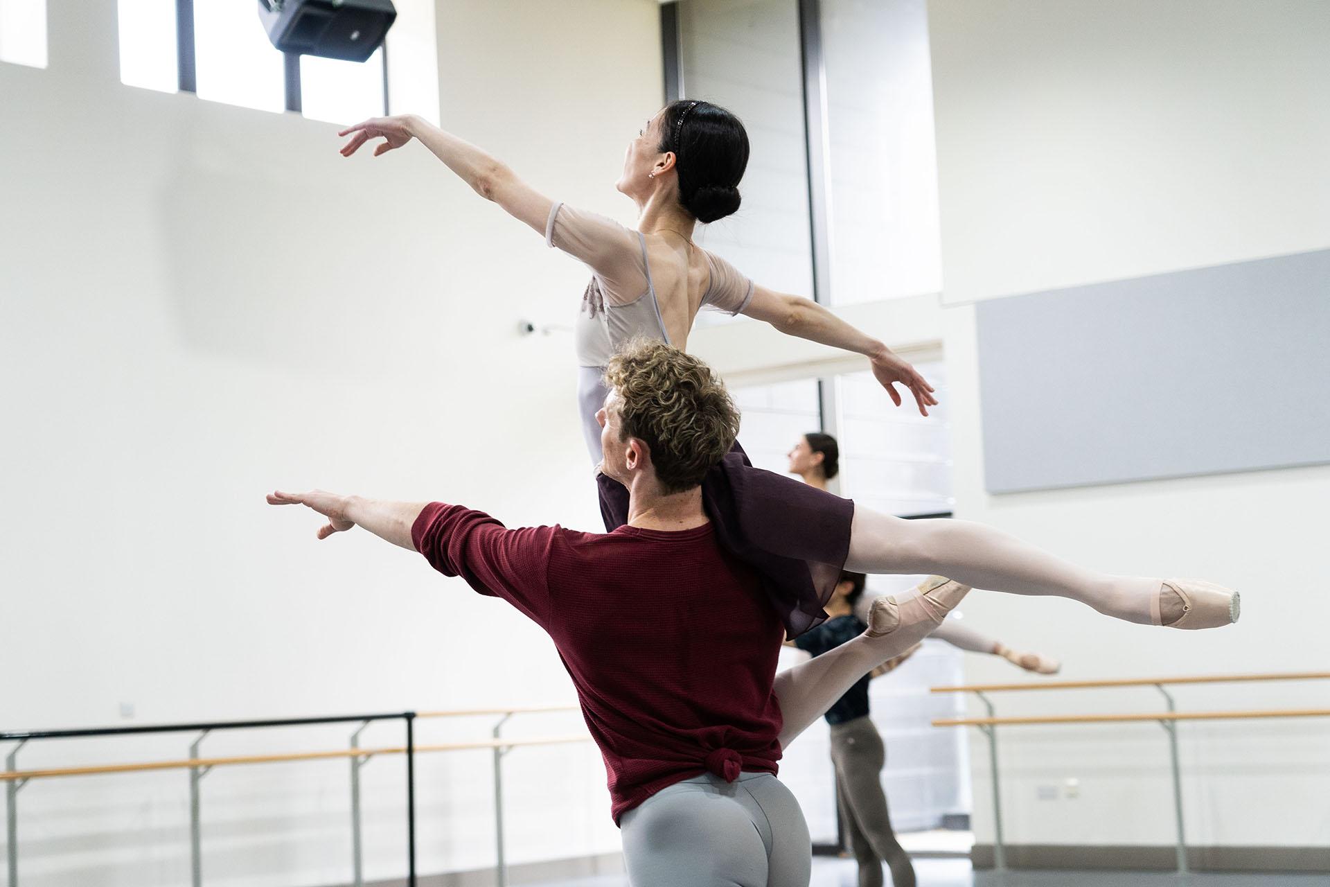 Dancer in a deep red top lifts another on his shoulder, the lifted person gracefully arches her back up, extends a hand in front of her and one leg behind