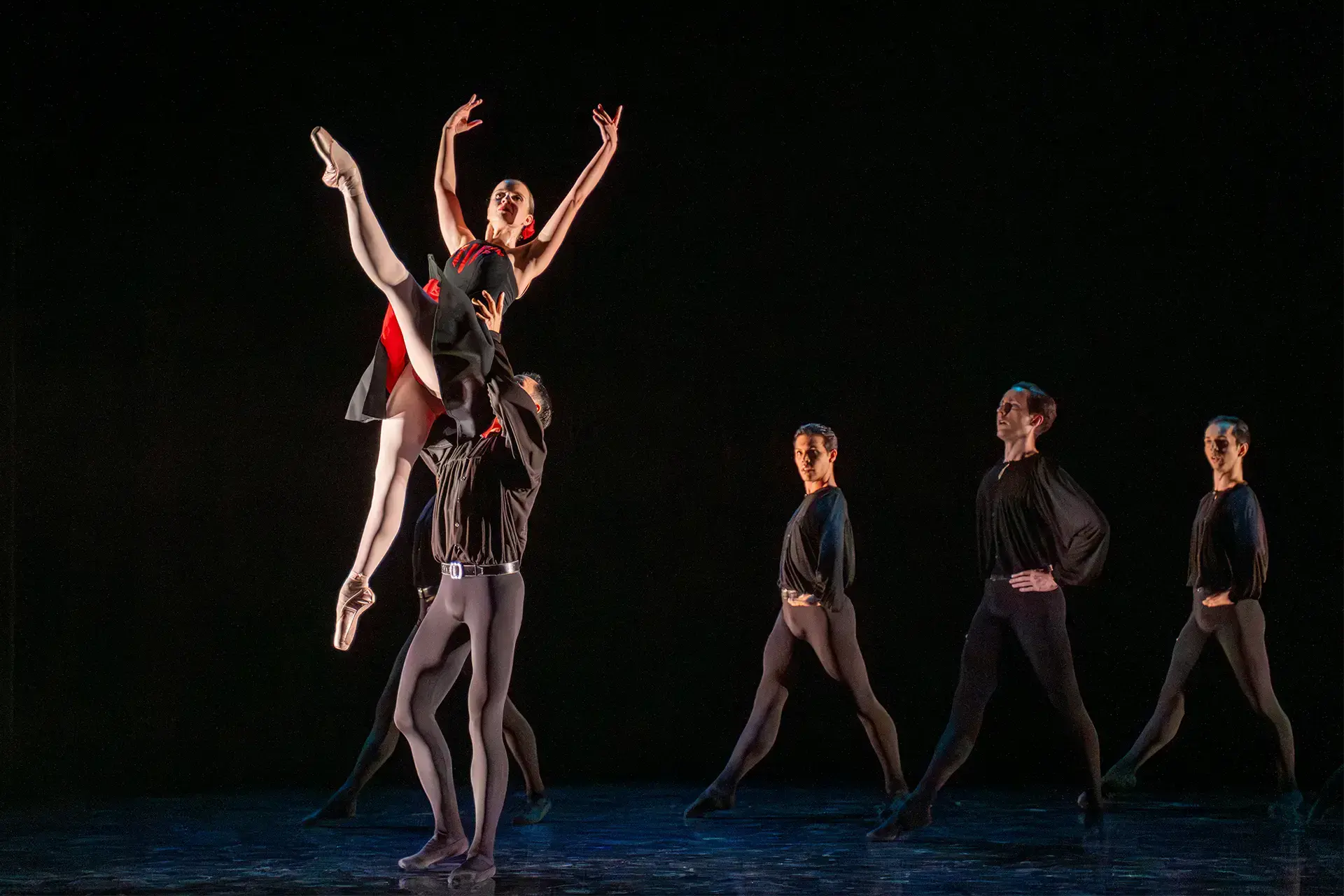 Dancer is lifted by the waist by a man wearing all black with three others watching