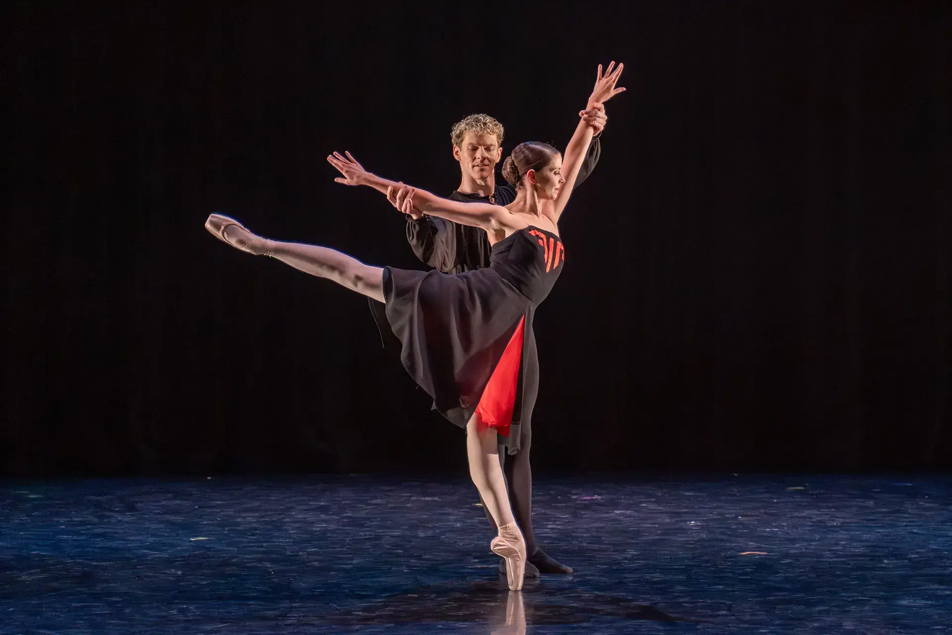 Dancer in a black dress with red accents stands arabesque supported by another dancer