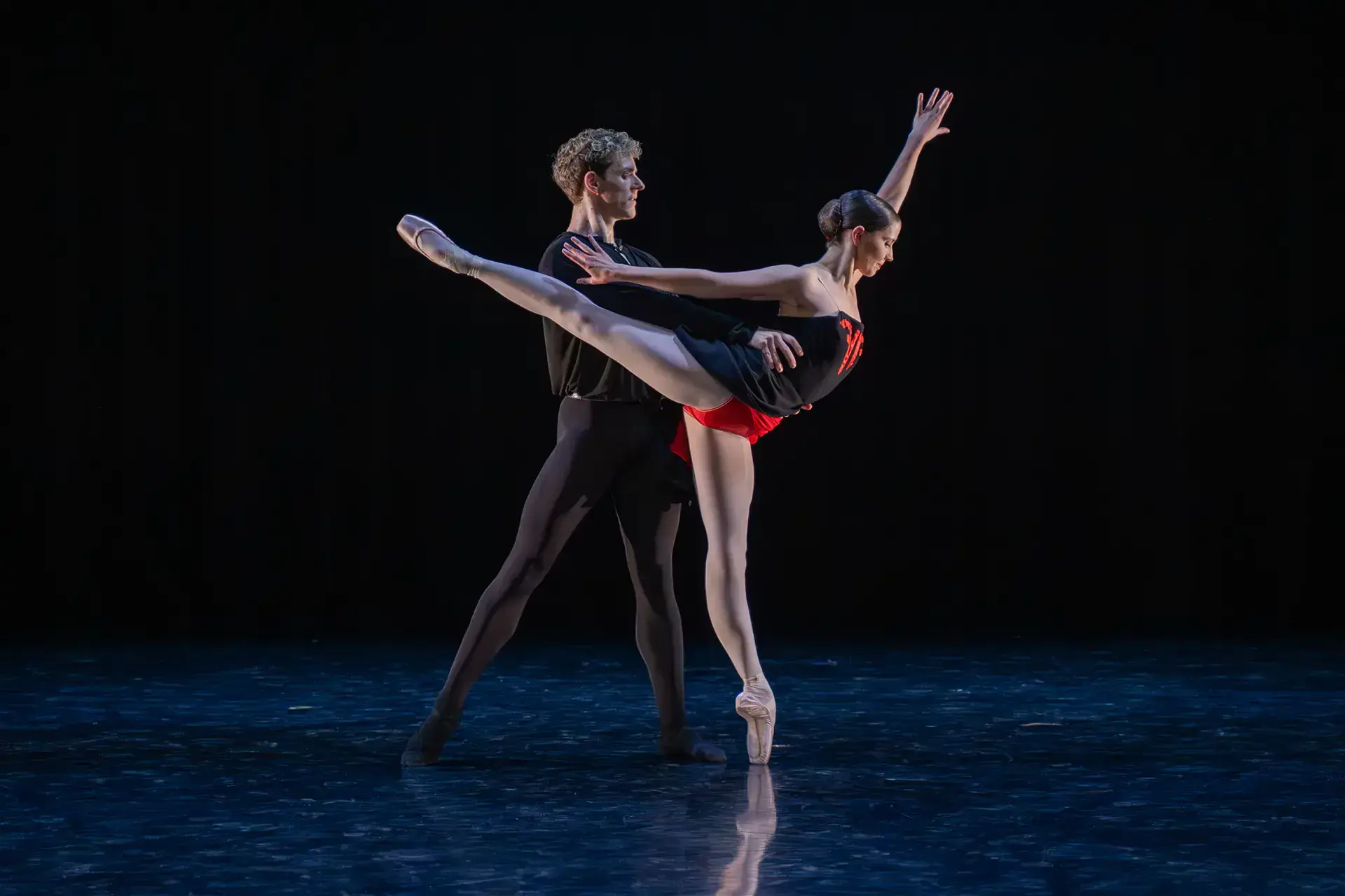 Dancer in a black dress with red accents stands arabesque supported by another dancer
