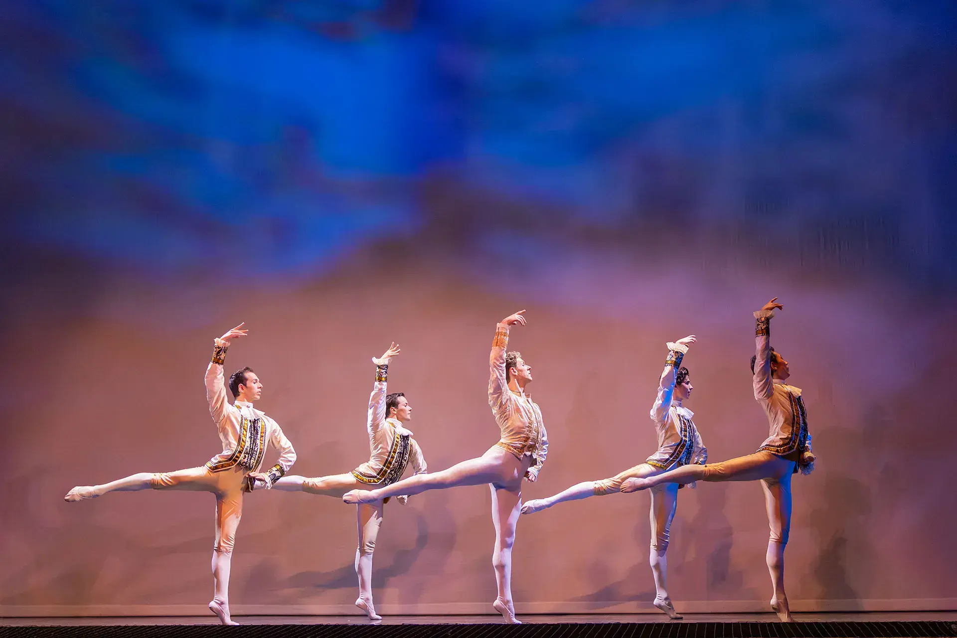 Five male dancers in white and bodies in sync, standing straight up on one foot, one hand on hip, the other arm high in the air