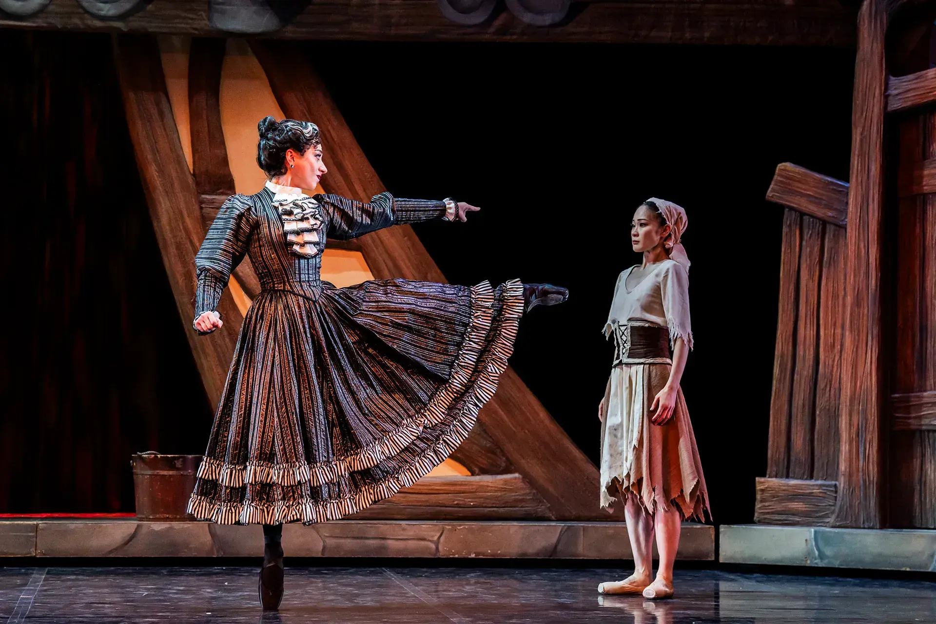 The stepmother, imperiously dressed and in black pointe shoes, point aggressively at Cinderella, who looks meek