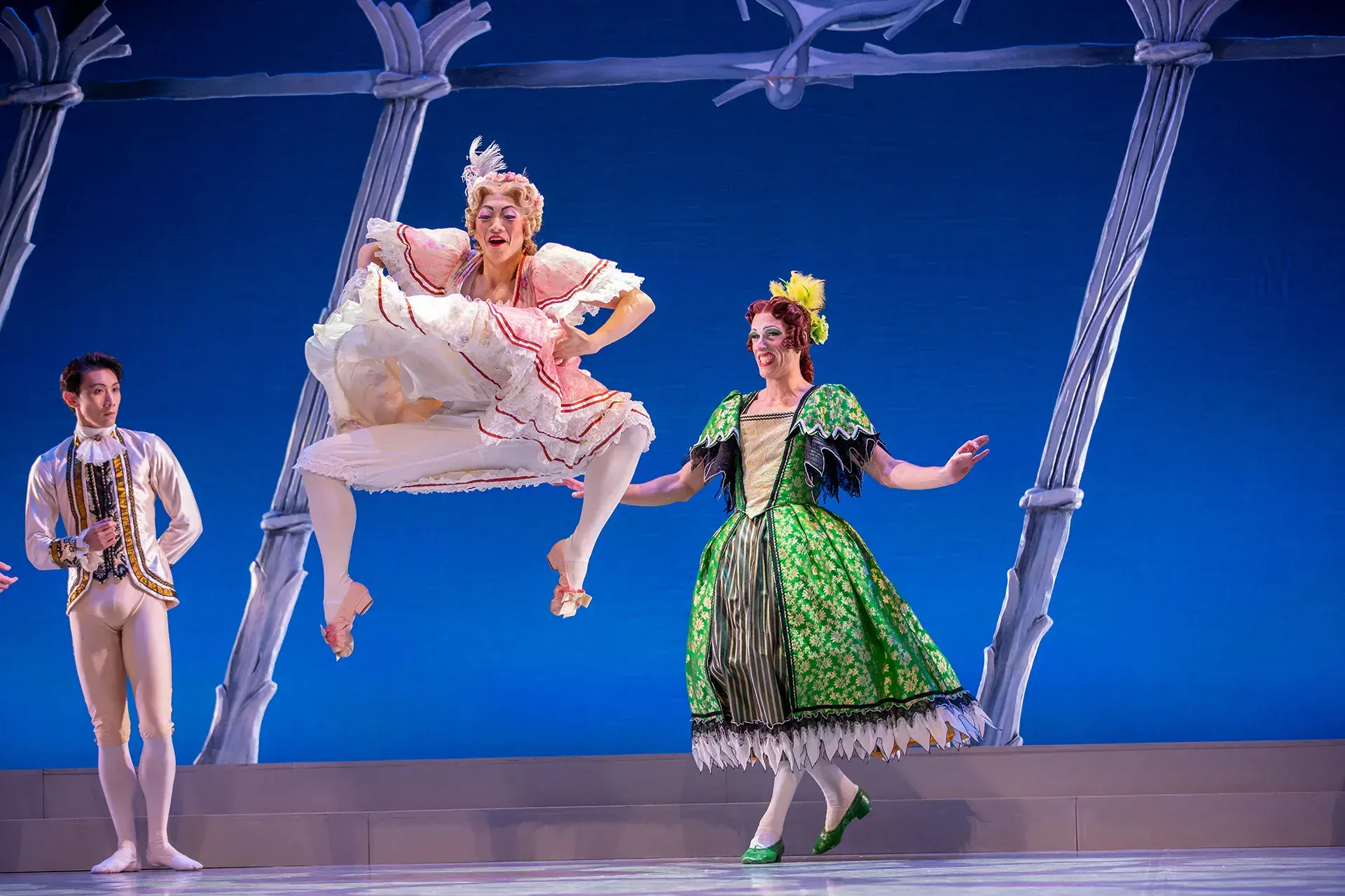 A wicked step-sister jumps high, lifting the skirt of her dress, her legs bent at the knees looking awkward