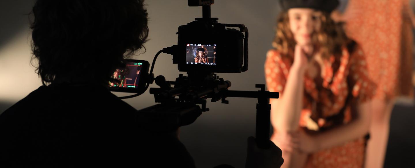 Behind the scenes at the filming of a dance film with students. We see the silhouette of the camera operator and the back of the camera, in the background we see a student looking down the camera, out of focus,