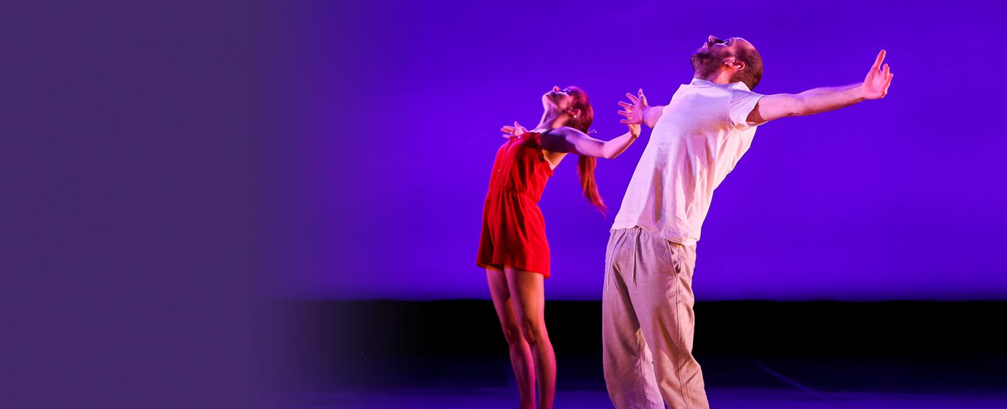 Two dancers perform at Expressions. They are stood at a diagonal to each other, with their arms outstretched and looking upwards. The floor and background is a bluey purple.