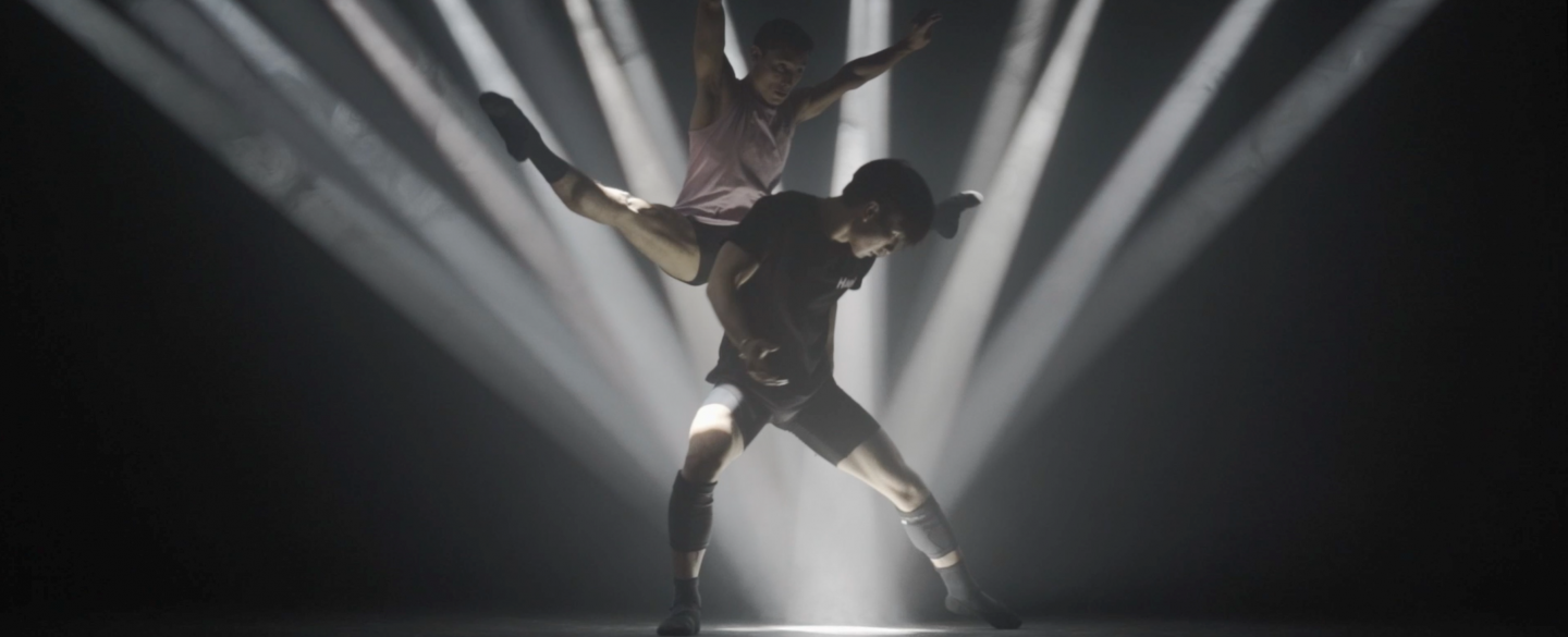 Beams of light focus on one spot. One dancer stands above it. A Second dancer leaps behind him, legs and arms spread apart.