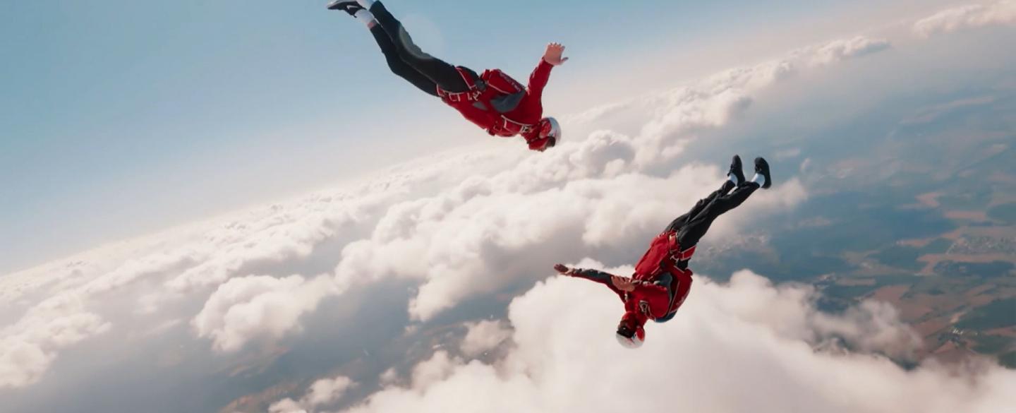 Two skydivers in red jackets and white helmets fall through the sky