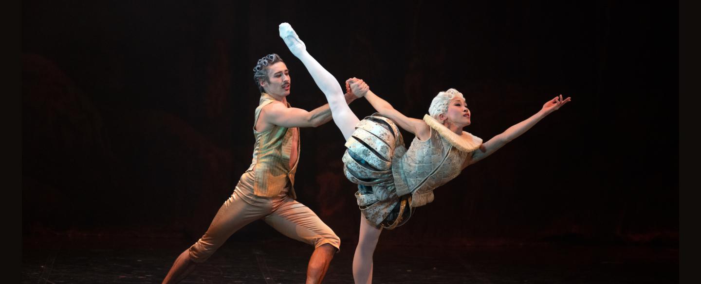 A female dancer in light 18th century costume reaches into the distance with her leg aloft, supported from behind by a male dancer in gold costume.