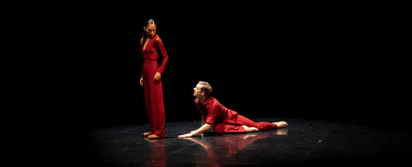 Two dancers dress in red, one is on the floor looking up at the other who is turned away but sees him over her shoulder