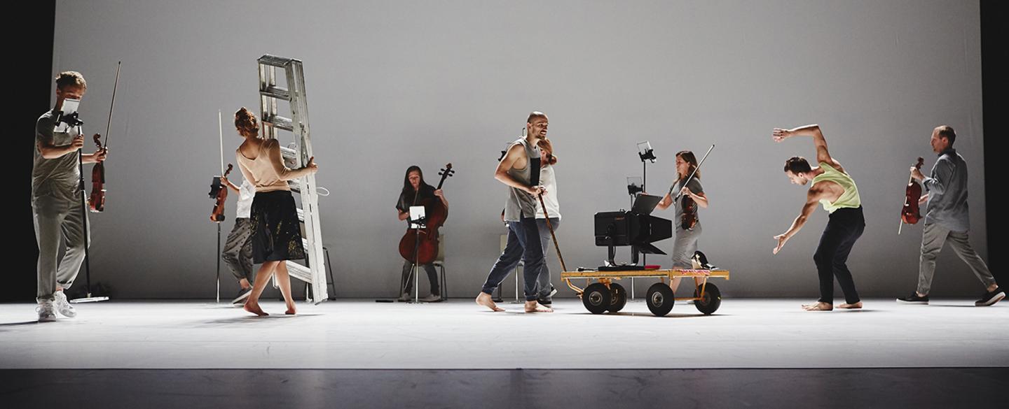 A group photo of a number of people on stage, one carrying a stepladder, another pushing a trolley, a third dancing, more people carrying and preparing stringed instruments