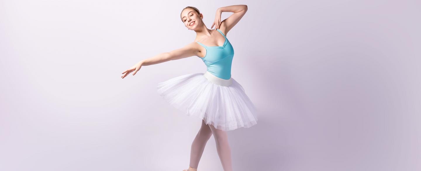 Student in blue leotard and white tutu stood on pointe with one arm outstretched