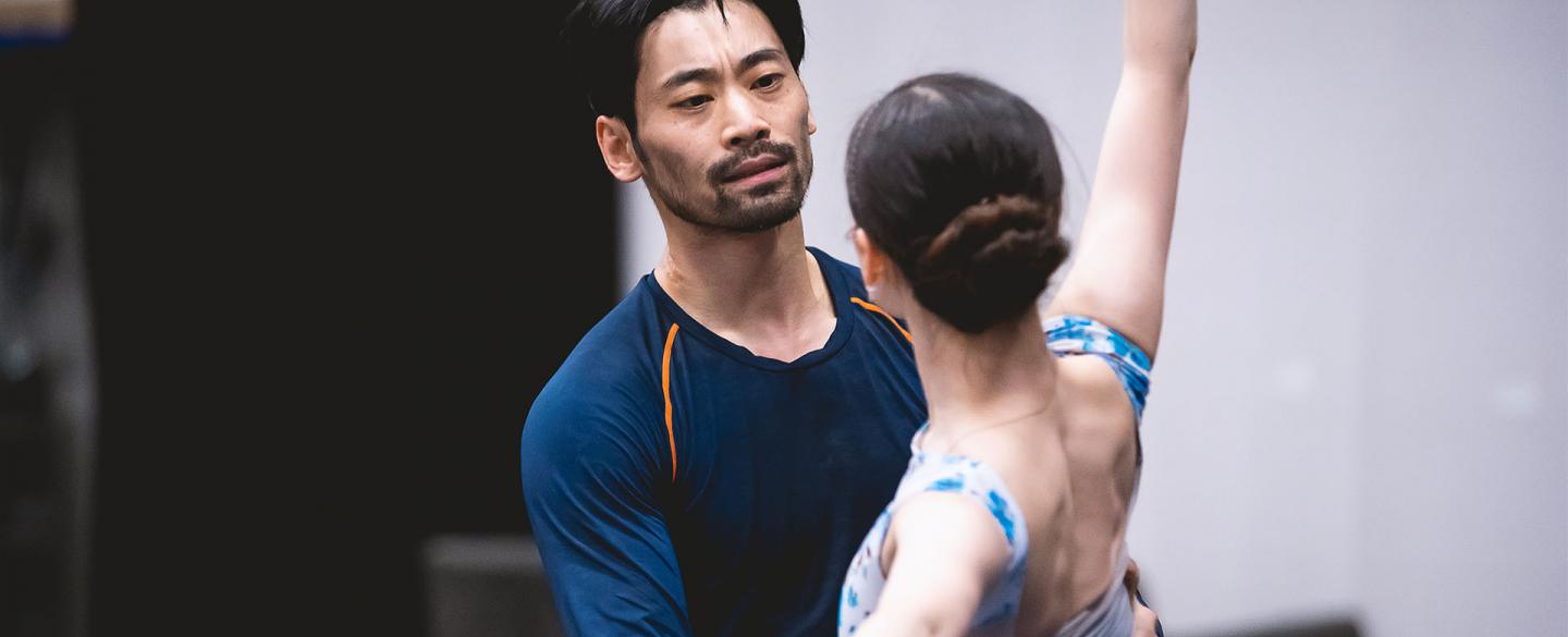 Dancers in rehearsal, one which one arm stretched abover her head and the other behind her, while the other holds her by the waist