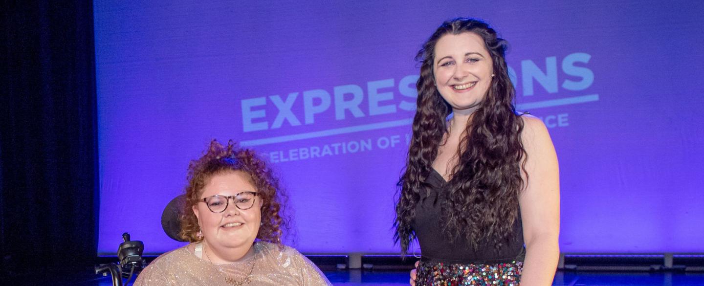 Two presenters are on stage at expressions. The woman on the left is in a wheelchair wearing a sparkly gold dress and red hair. The woman on the right is stood up with a walking stick, wearing a black top, gold sequin skirt and long black hair.
