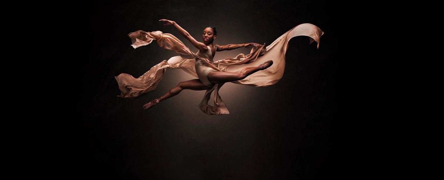 Dancer leaping across the frame while looking directly at the viewer, wearing peach and with fabrics flowing in the wind