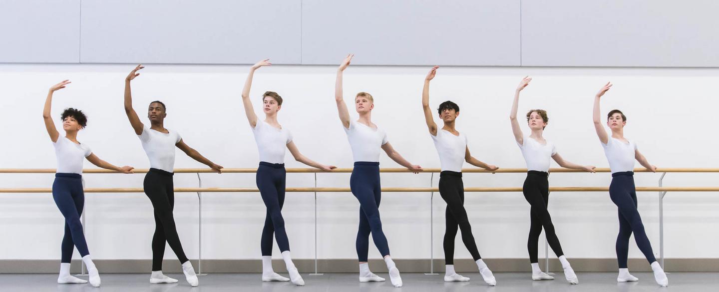 Seven male dancers stood at the bar with one arm raised 