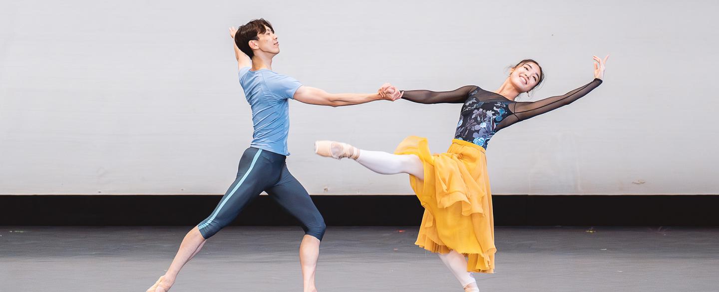 Two smiling dancers, one supporting the other with an outstretched hand, and the other, in a yellow dress and dark patterned top, stands on one leg, her other stretched out in front of her