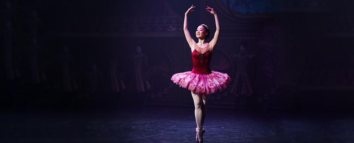 Dancer en pointe, wearing a pink top and tutu as the Sugarplum Fairy with her arms raised above her head