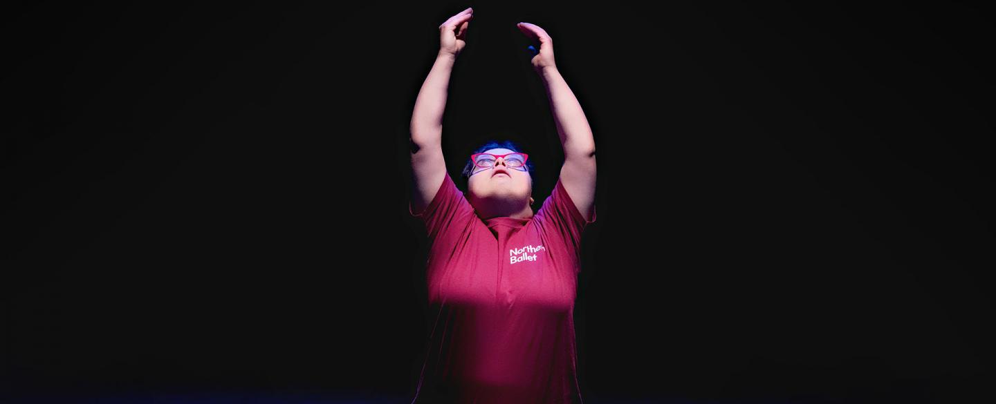 PErformer in a red T-Shirt with a darkened background looks up and reaches both of her arms straight up