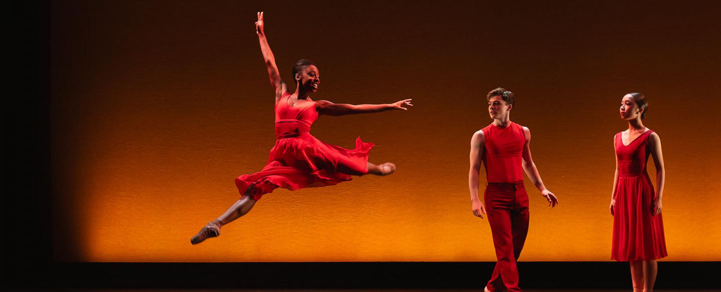 Female dancer wearing a red dress leap high with an arm stretched above her while two dancers dressed in red look on