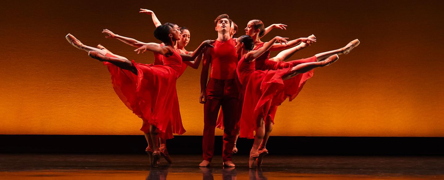 A male dancer in red is standing with arms by his side as female dancers also in red, stand arabesque around him