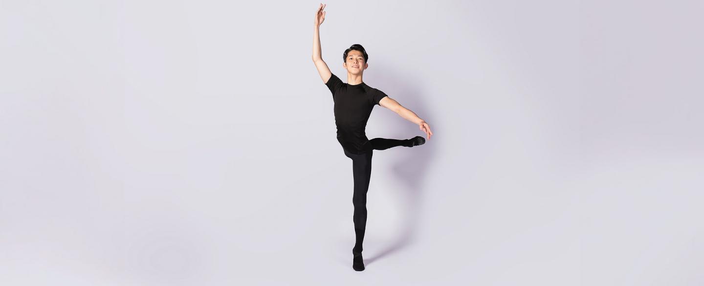 Dancer smiling at the camera, dressed in black, standing on one foot, his other leg raised behind him. His arms are in fourth position, one above his head, the other slightly raised to the side.