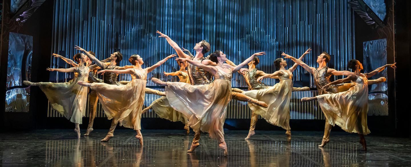 A dozen dancers in gold and white dance in unison, each stood on one foot, the other leg in arabesque behind them