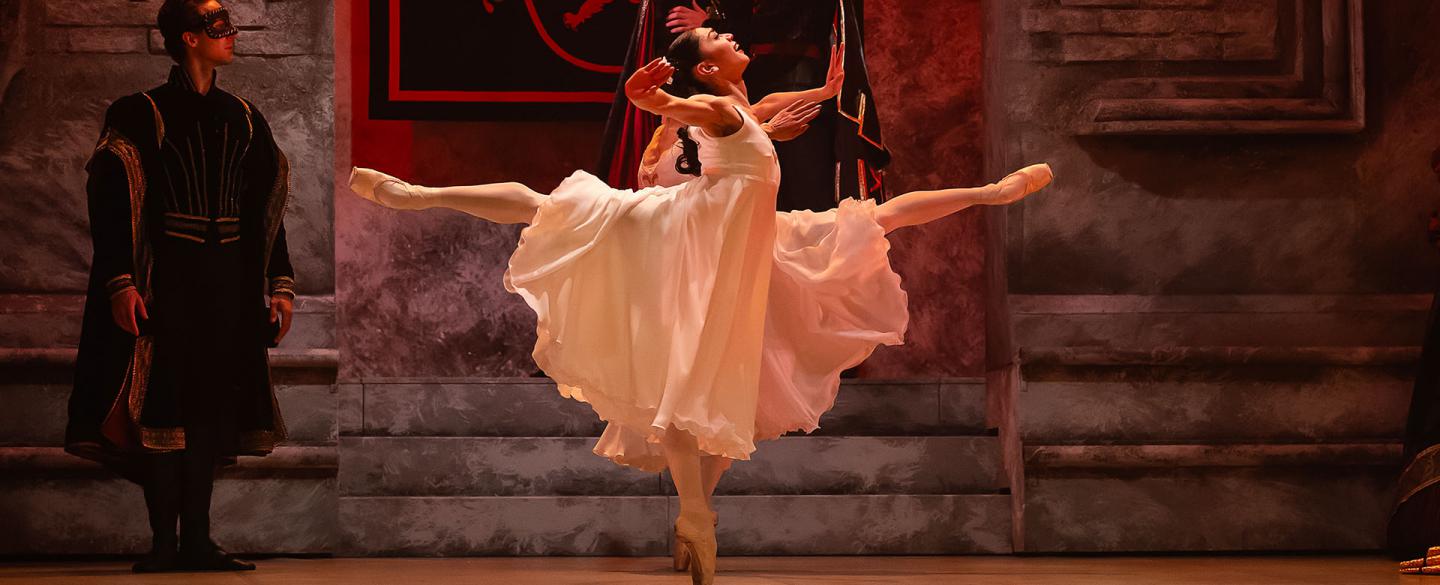 Dancer dressed in a white flowing dress dances elegantly, one leg raised behind her perpendicular to the floor while en pointe with the other