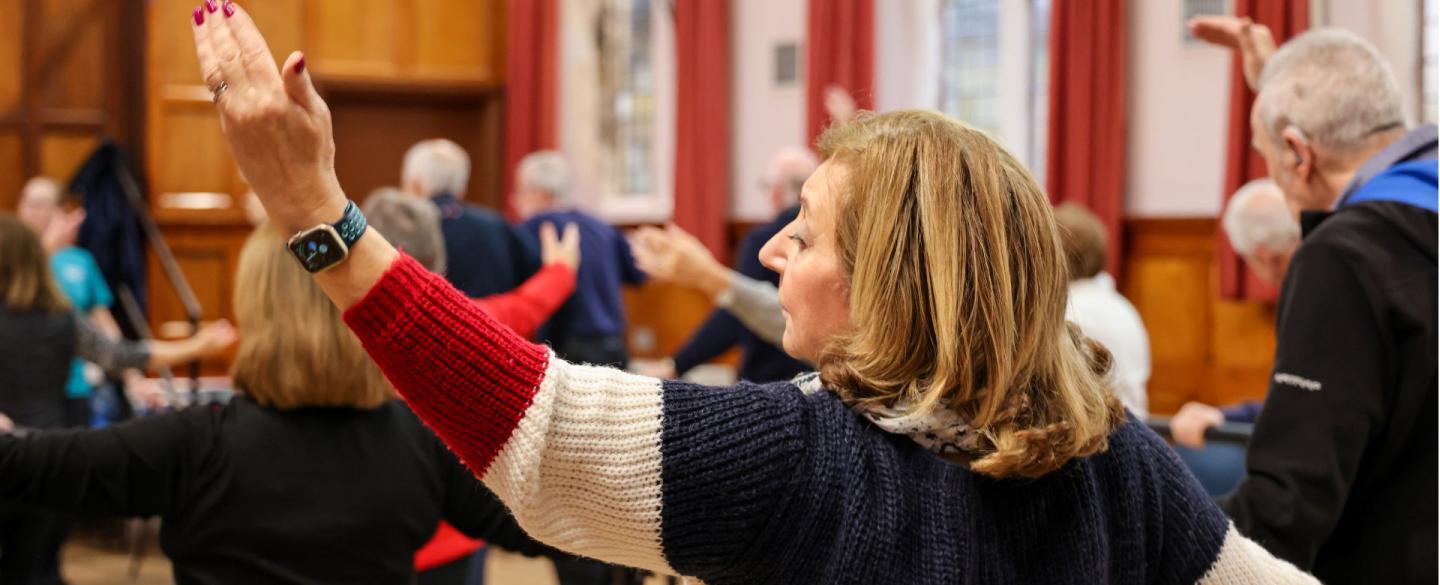 A Dance for Parkinsons class where many attendees are seen raising their arm at a barre