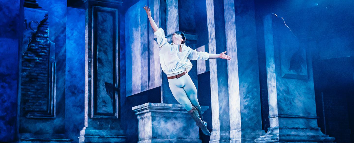 Dancer leaping across the stage, his legs together, one arm raise triumphantly above him, bathed in blue light